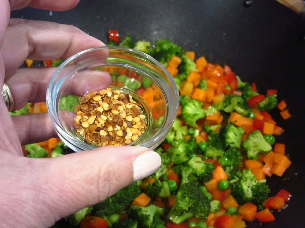 Red pepper flakes in a small glass bowl held over a wok with chopped vegetables.