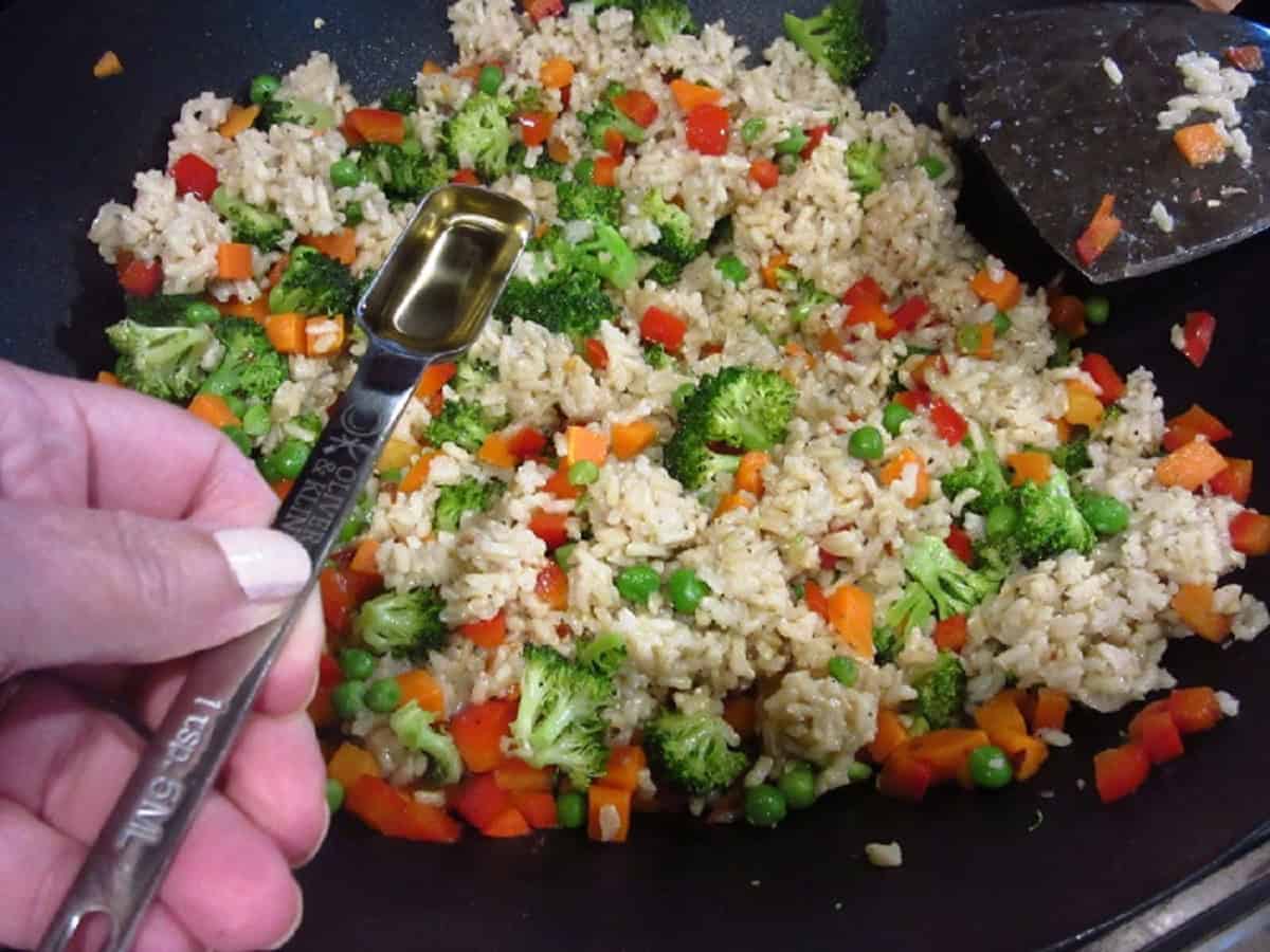 A measuring spoon with vinegar in front of stir-fried brown rice in a wok.