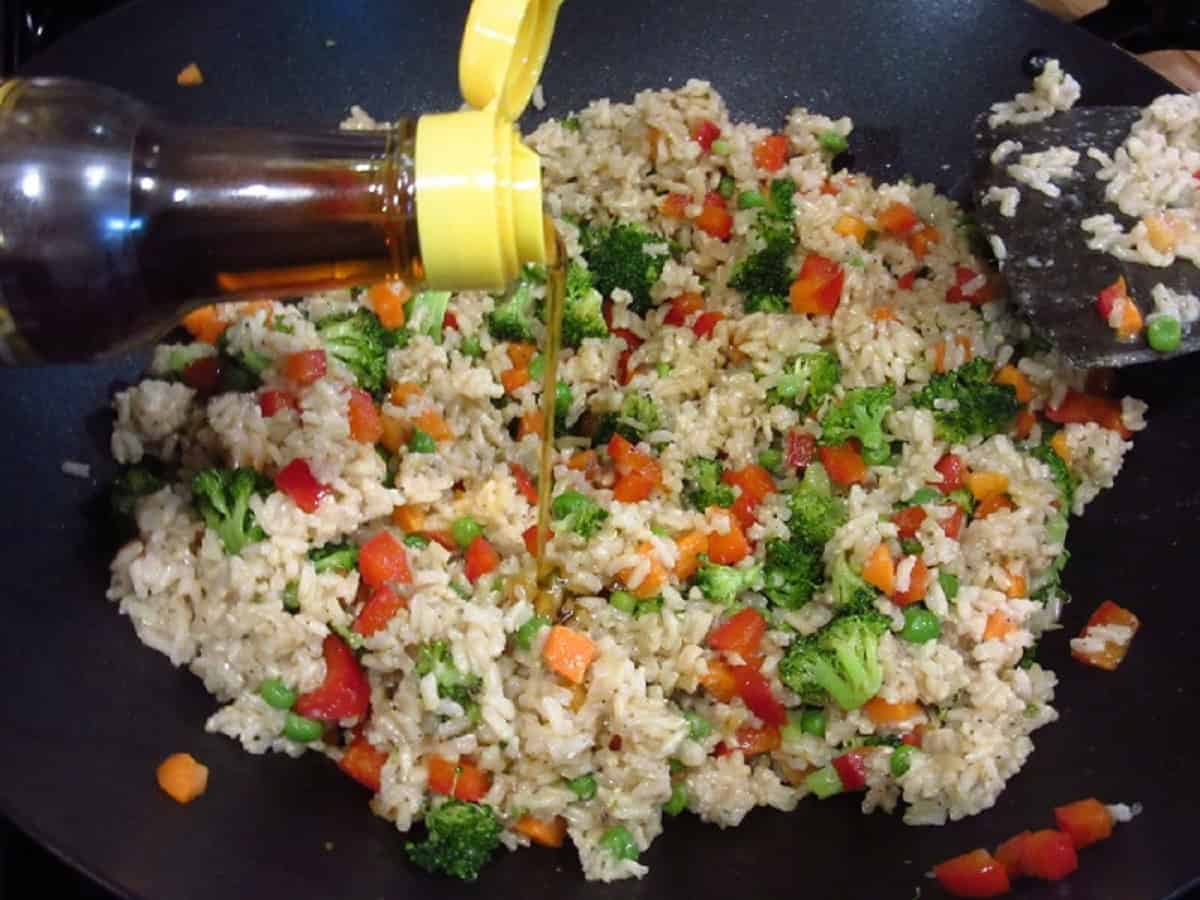 Sesame oil being poured into a wok full of stir-fried brown rice.
