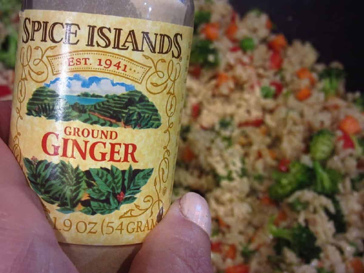 A jar of ground ginger being held in front of stir-fried brown rice.