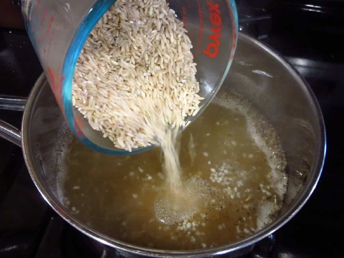 Uncooked brown rice being poured into a simmering saucepan.
