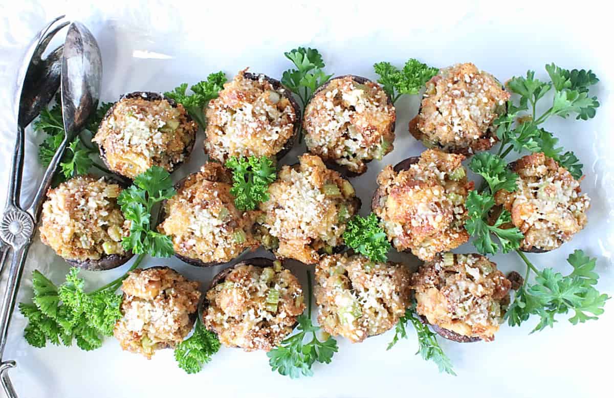 A white platter filled with stuffing stuffed cremini mushroom caps along with fresh parsley and a pair of tongs.
