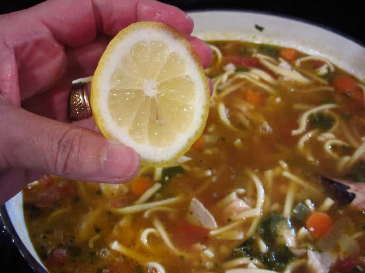 A hand holding a slice of lemon over a pot of chicken noodle soup.