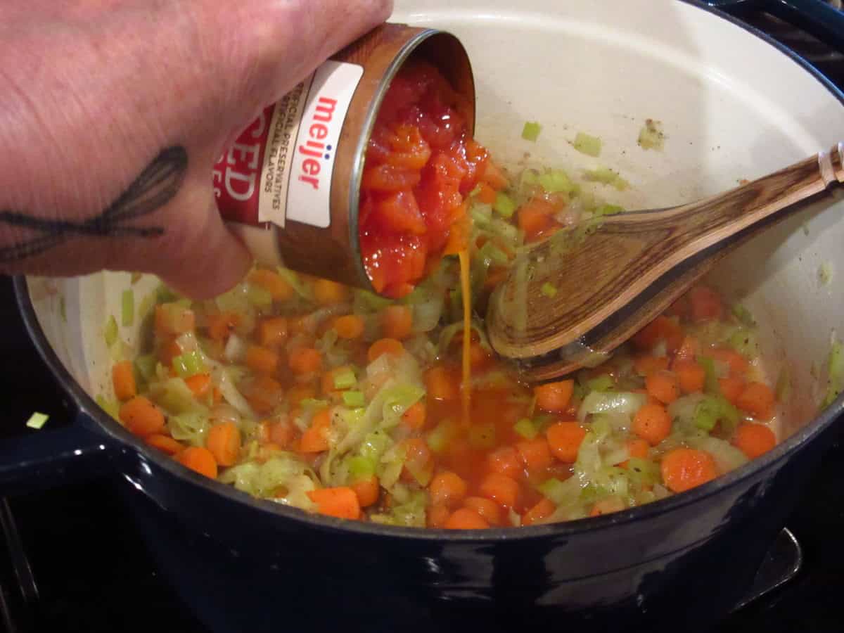 A can of diced tomatoes being added to a Dutch oven making chicken noodle soup.