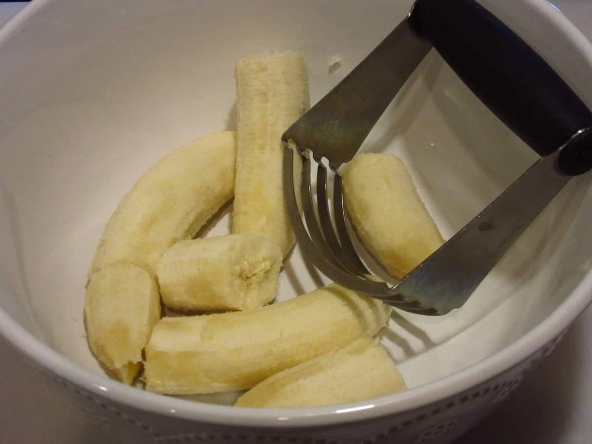 Unpeeled bananas in a white bowl with a pastry blender.