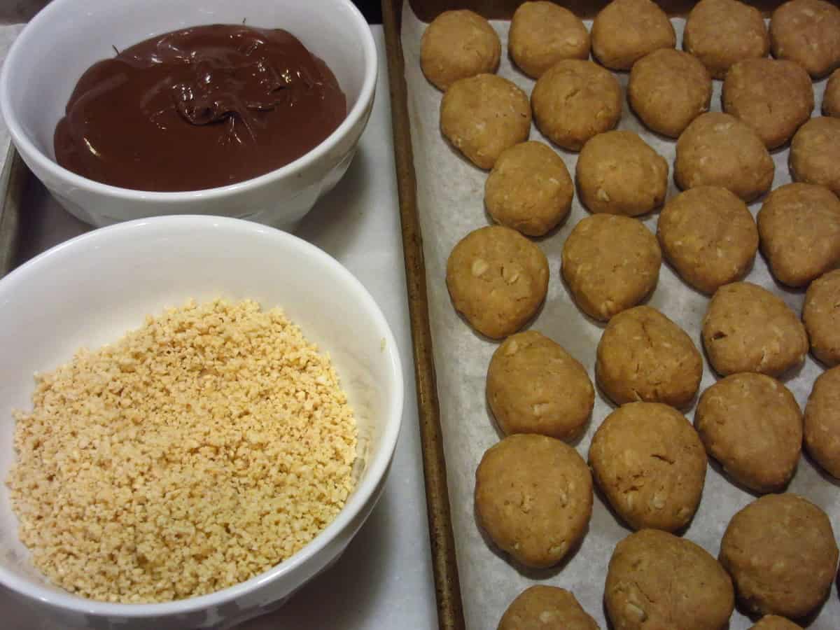 Chopped peanuts and melted chocolate in small bowls next to a cookie sheet filled with baked acorn cookies.