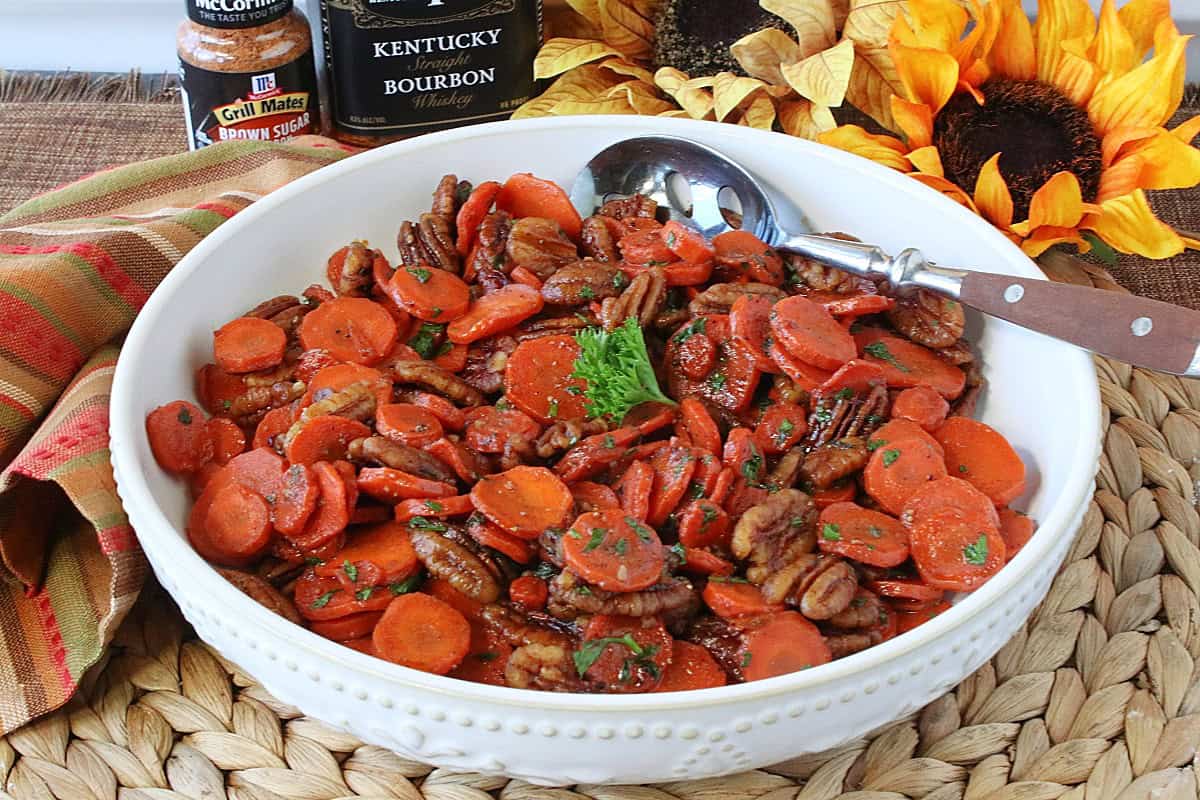An autumnal looking side dish of bourbon glazed carrots and pecans in a bowl with sunflowers in the background.