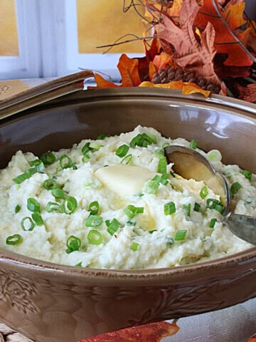 A tan casserole dish filled with Mashed Parsnips and Scallions along with melted butter and a spoon.