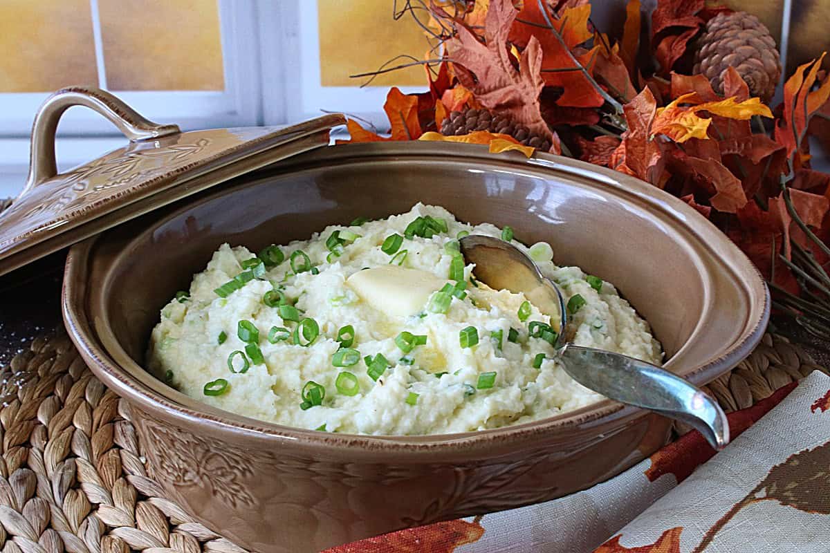 A fall scene with a casserole dish filled with Mashed Parsnips and Scallions with leaves in the background.