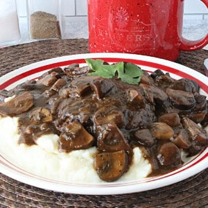 A red and white plate with a serving of ground beef salisbury steak covered in a deep brown mushroom gravy.