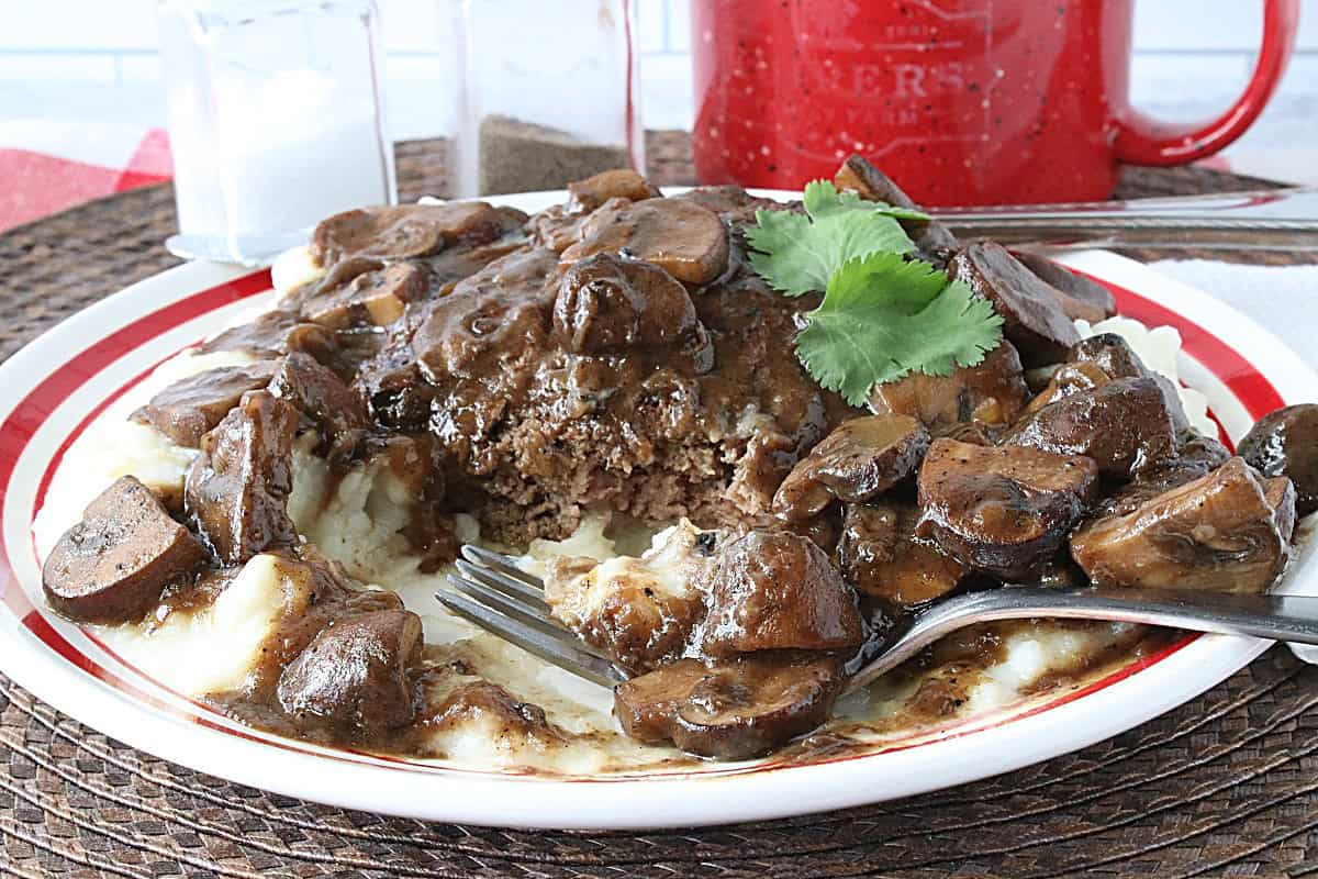 A red and white plate with a serving of Salisbury Steak and mashed potatoes and mushrooms.
