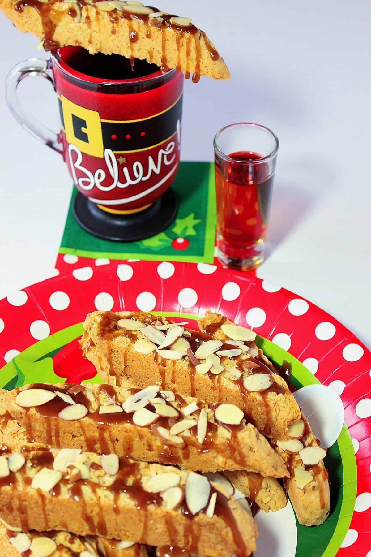 A Christmas plate piled high with almond coconut biscotti and a caramel drizzle along with a shot glass and cup in the background.