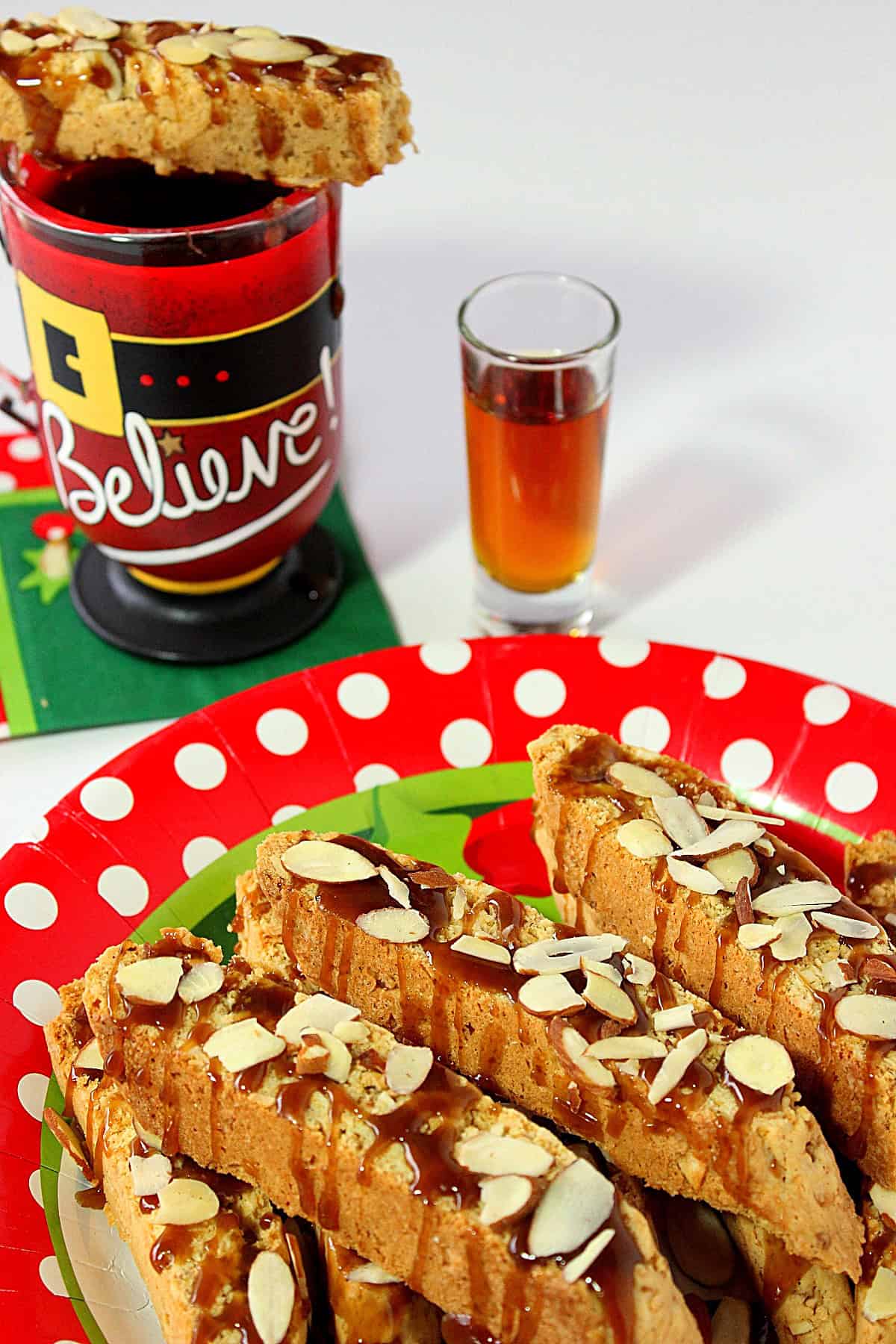 A shot of amaretto along with a Santa mug of coffee next to a plate of Amaretto Coconut Biscotti.