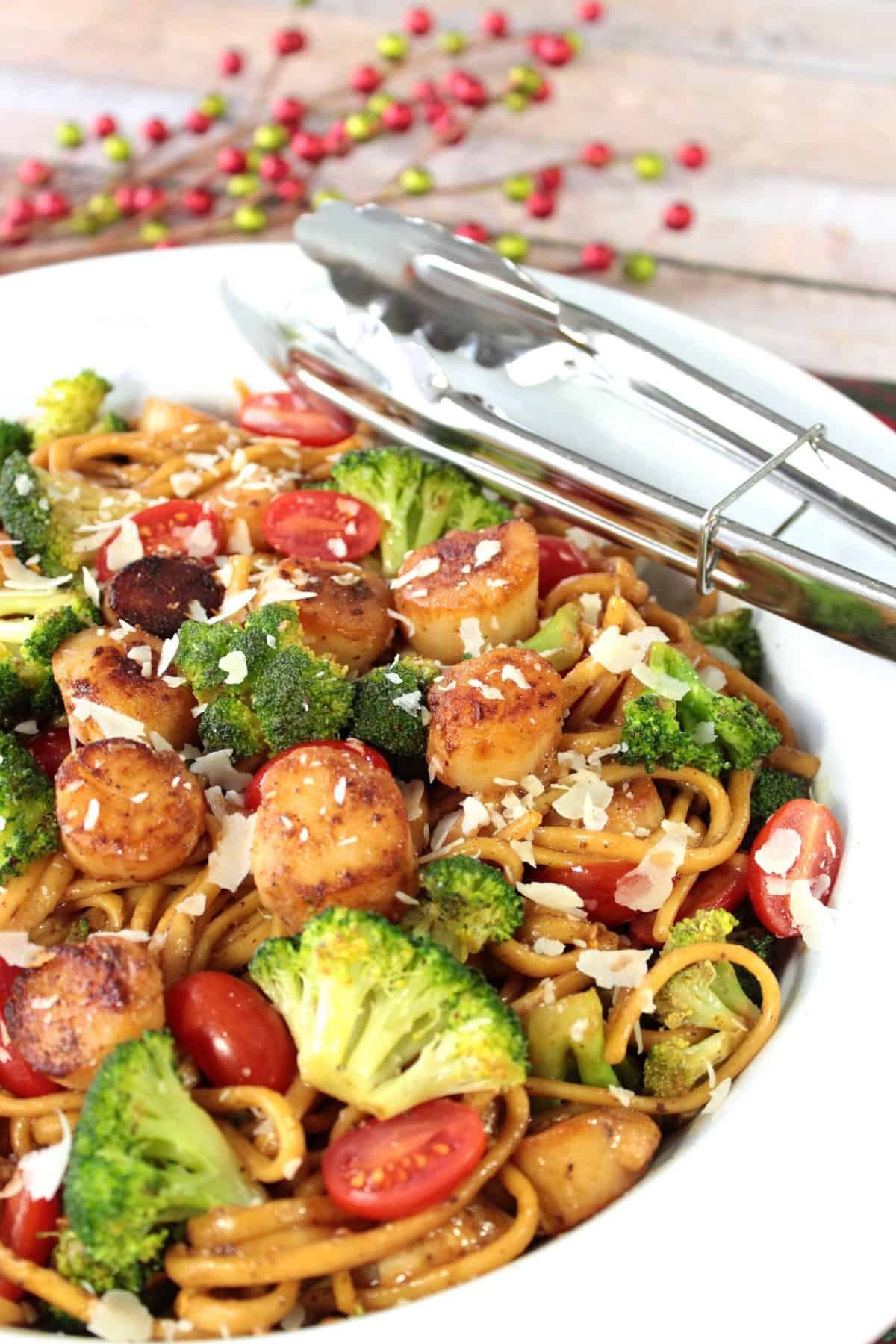 Tongs on the side of a bowl filled with pasta and scallops.