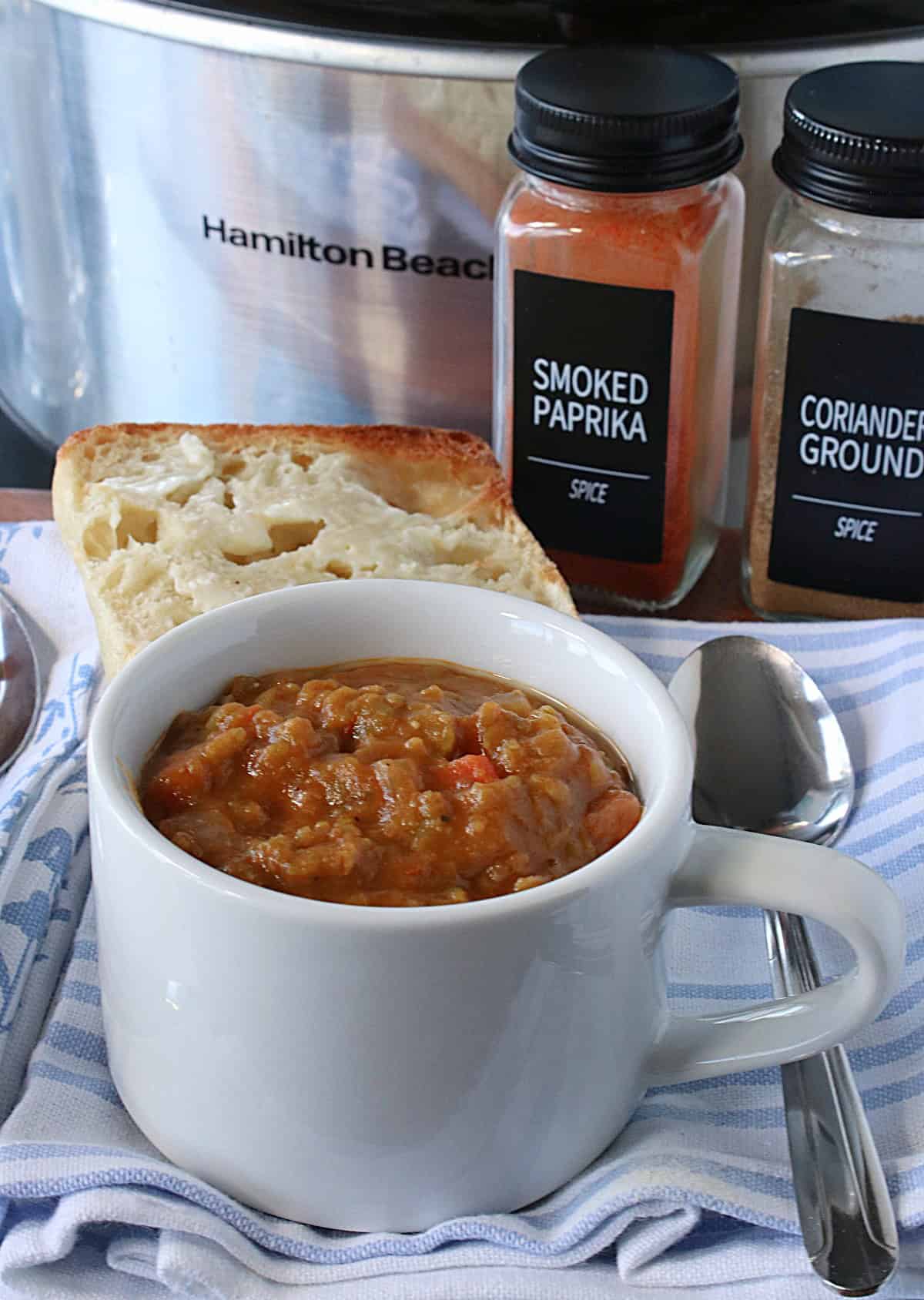 Two spice jars in the background and a cup of slow cooker split pea soup with ham in the foreground.