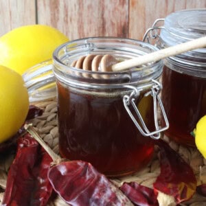 A jar of hot honey with lemons and dried peppers surrounding it.