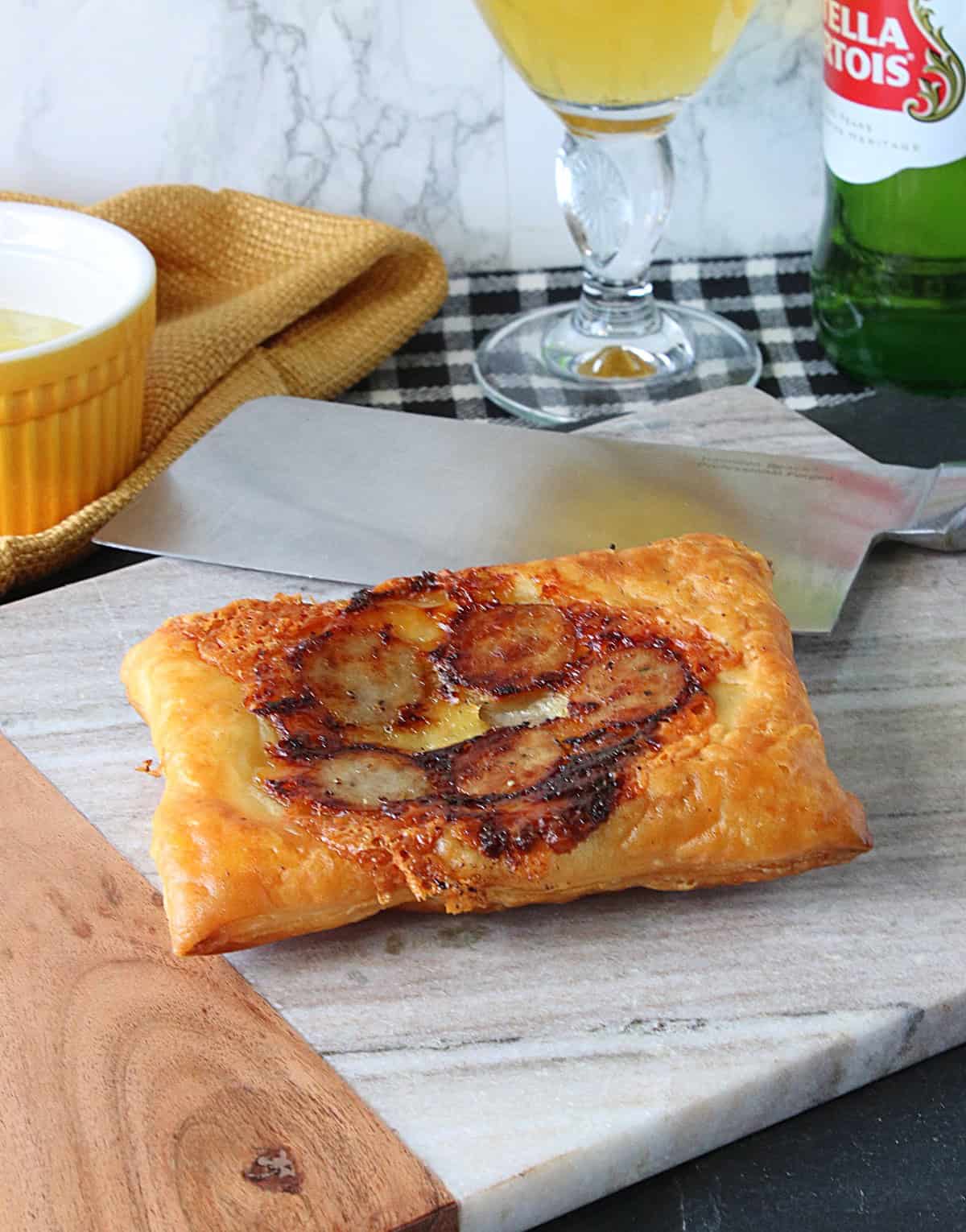A flaky pastry with cooked bratwurst and cheese on a marble tray.