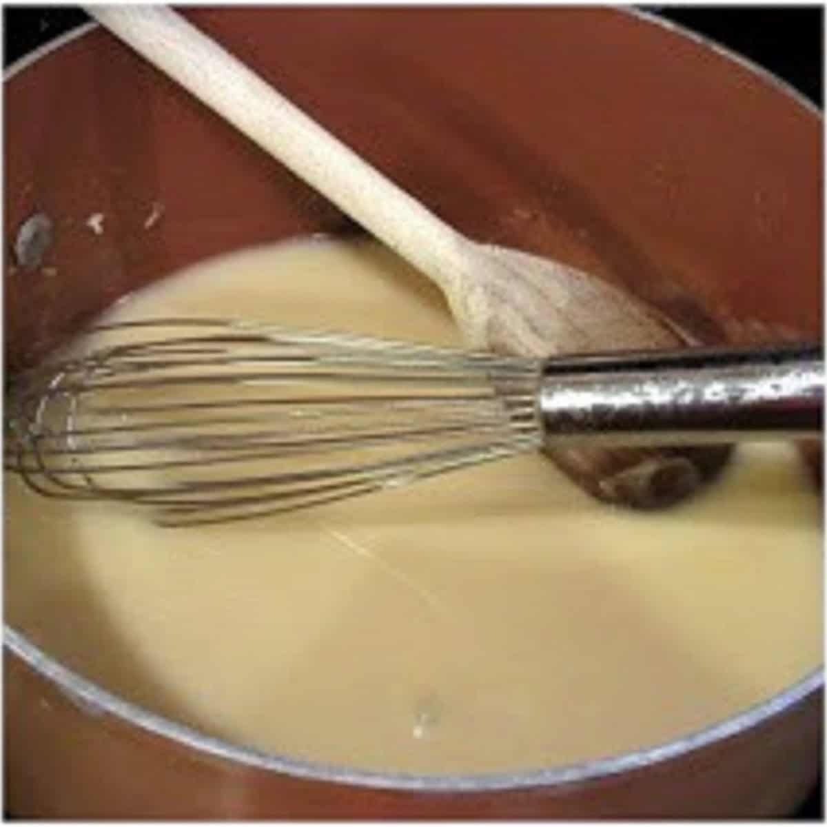 A whisk and wooden spoon in a saucepan with a chicken sauce.