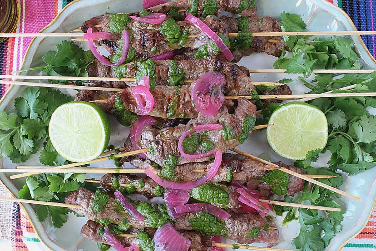 A pretty platter filled with Grilled Beef Flank Steak Skewers topped with chimichurri sauce and red onion.