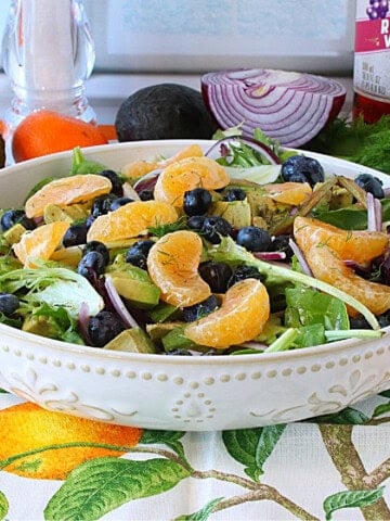A pretty white salad bowl with a lettuce salad with oranges, avocado, blueberries and fennel inside.
