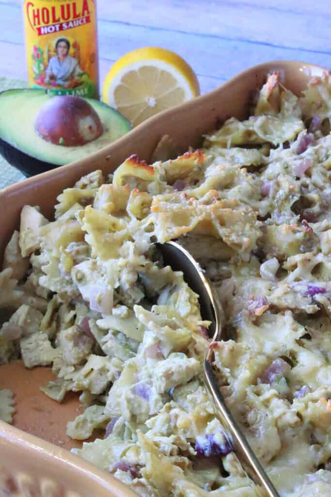 A serving spoon in a casserole dish of bow tie pasta, chicken, artichokes, and avocados.