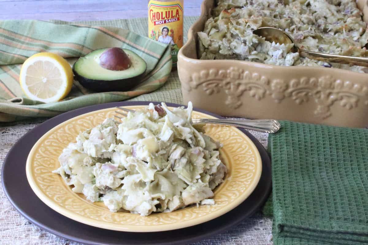 A yellow plate with a serving of chicken artichoke pasta in the foreground and the casserole dish in the background.