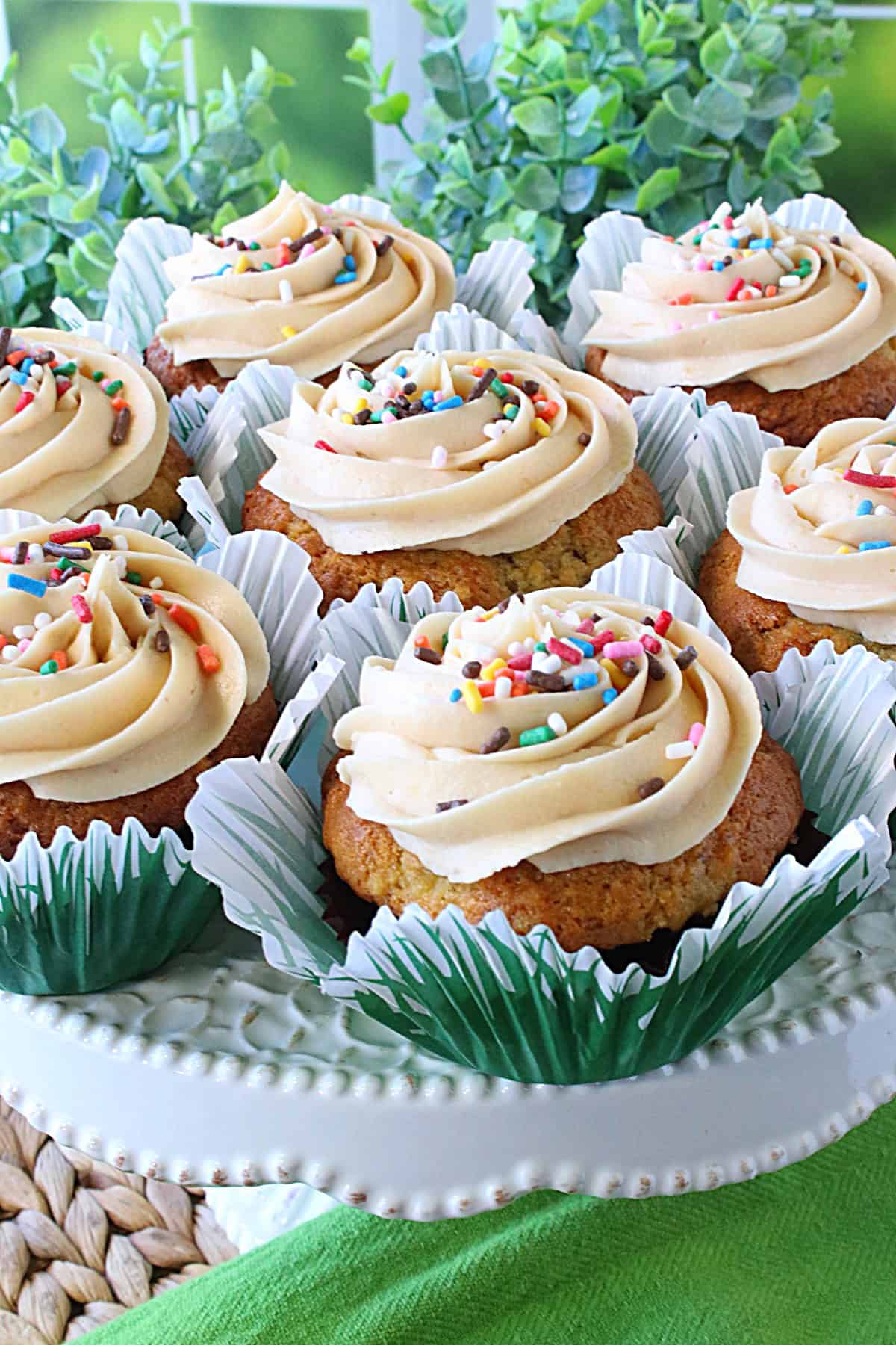 Sprinkles and swirly frosting on banana cupcakes on a cake plate.
