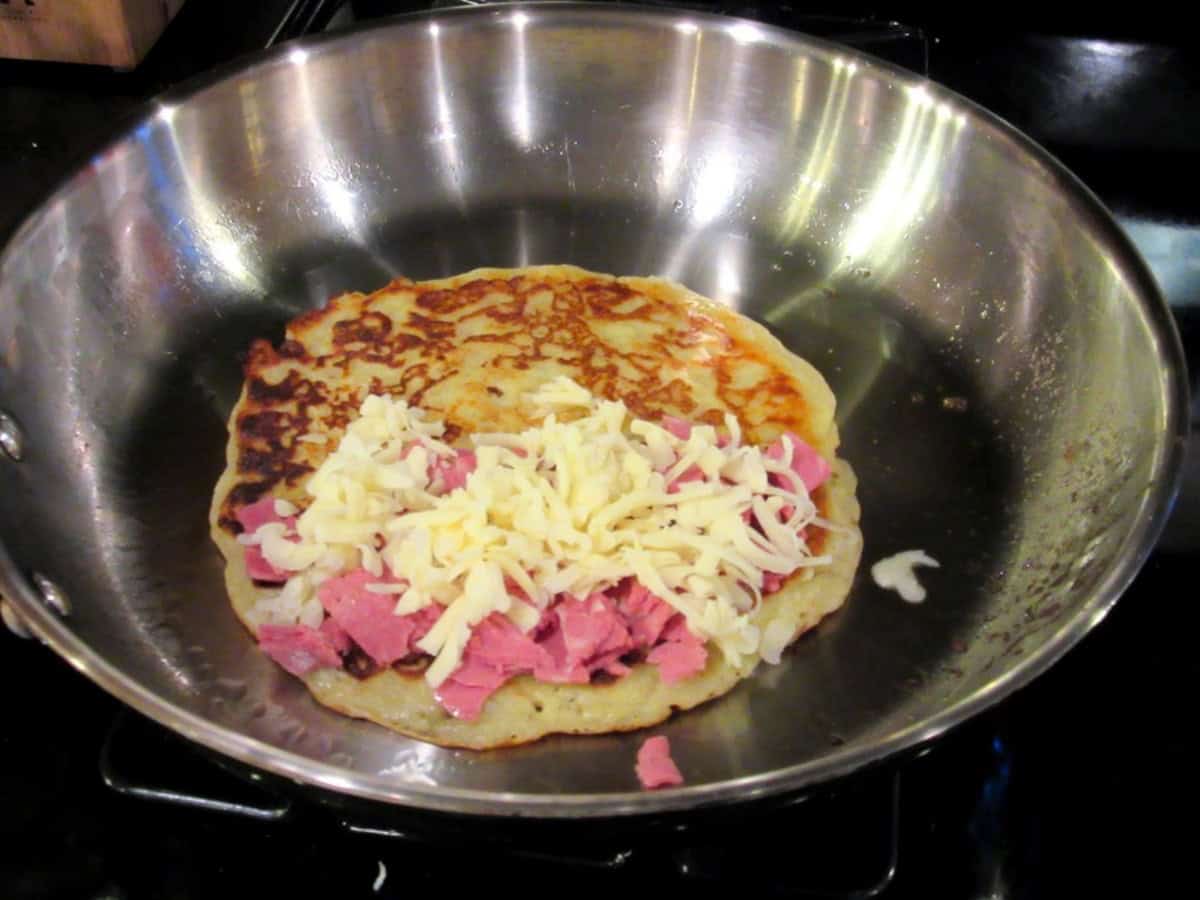 A potato pancake in a skillet topped with corned beef and shredded Swiss cheese.