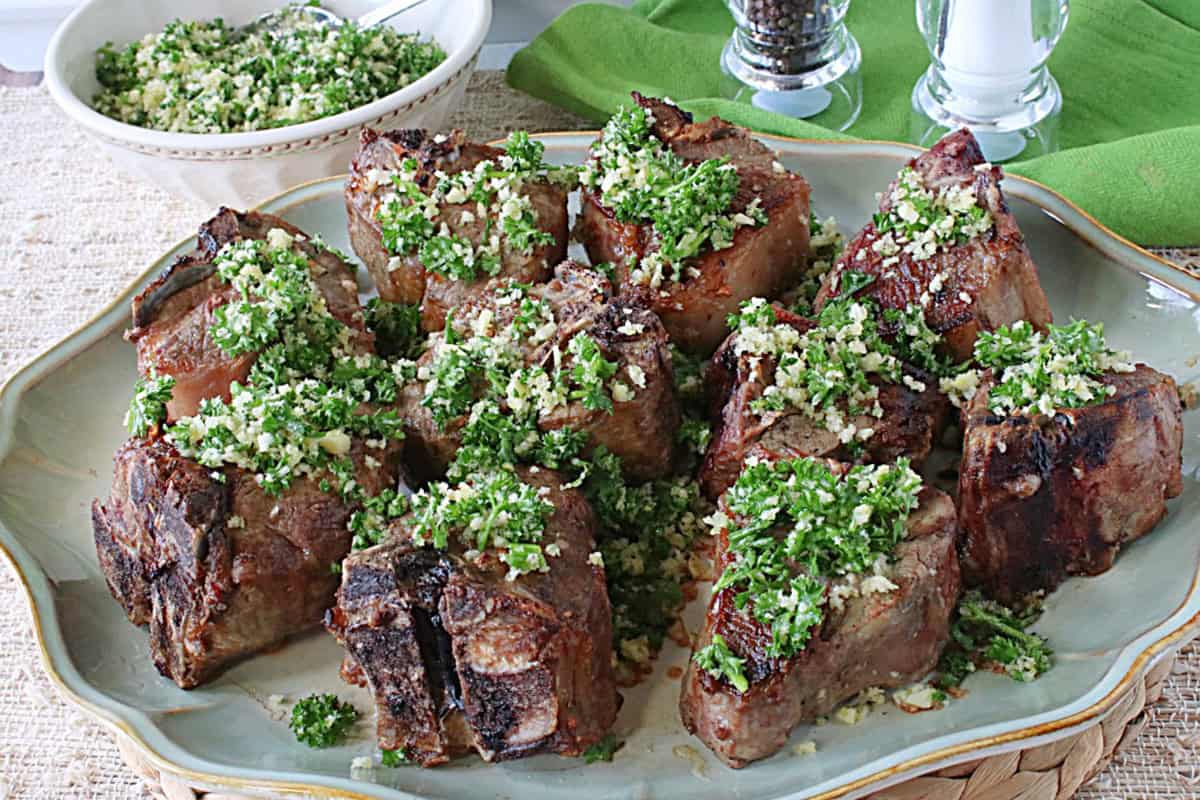 Ten cooked lamb chops on a platter all topped with persillade.