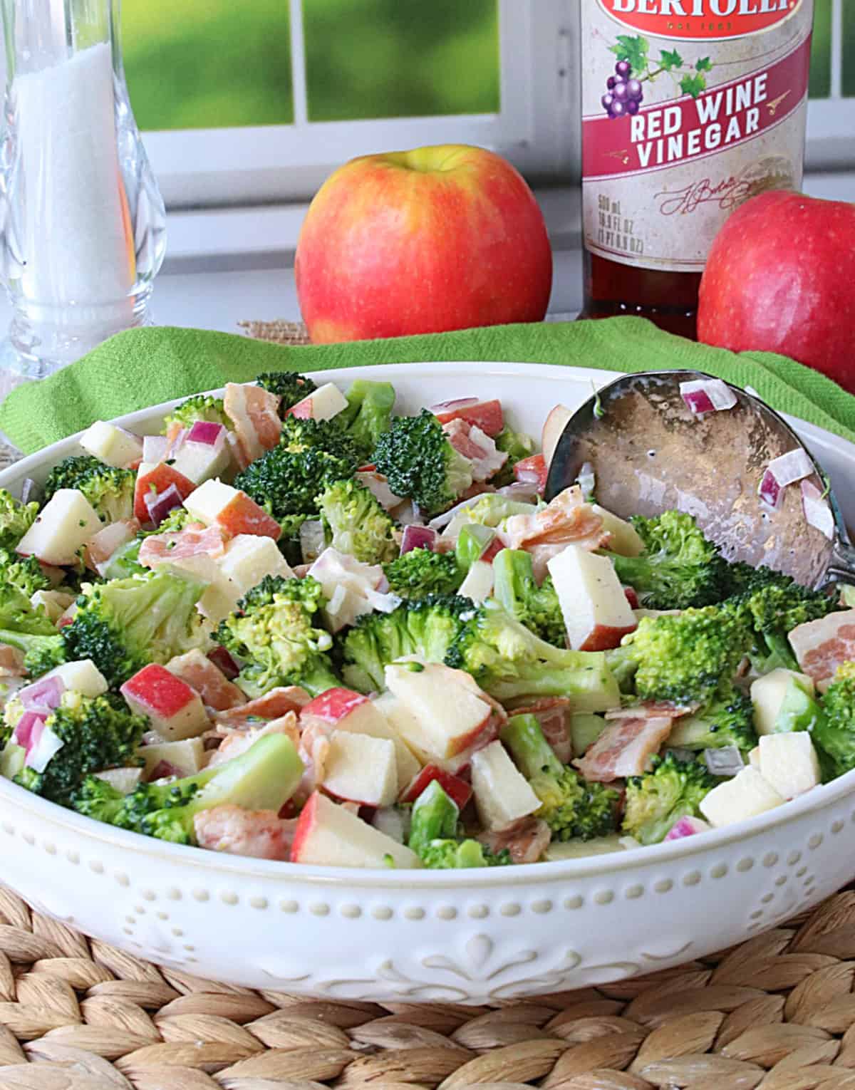 A pretty white bowl filled with broccoli and apple salad with red apples in the background.