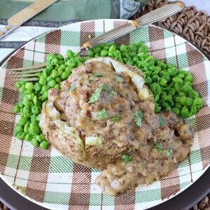 A cooked and crumbled bratwurst gravy over a baked potato on a plate with peas.