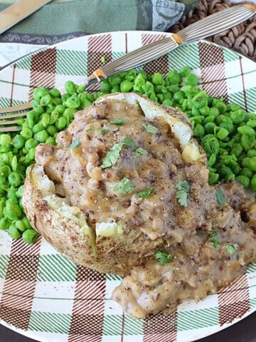 A cooked and crumbled bratwurst gravy over a baked potato on a plate with peas.