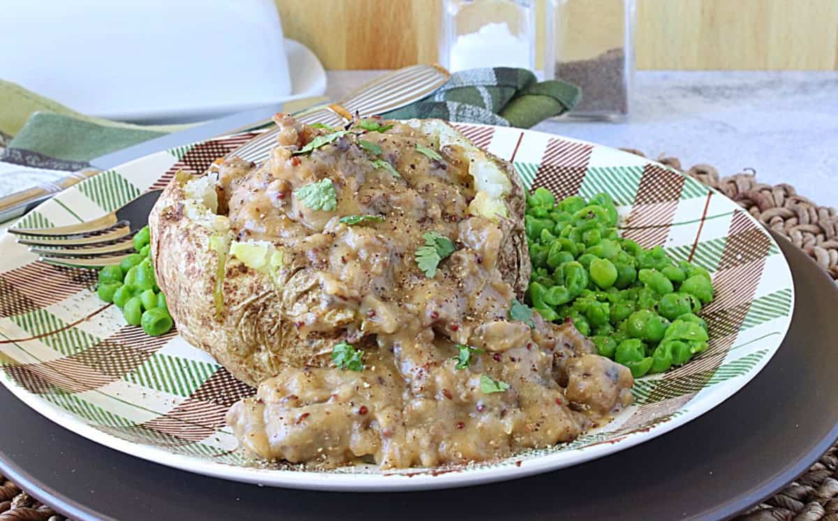 A baked potato topped with sausage gravy with mashed peas on the side.