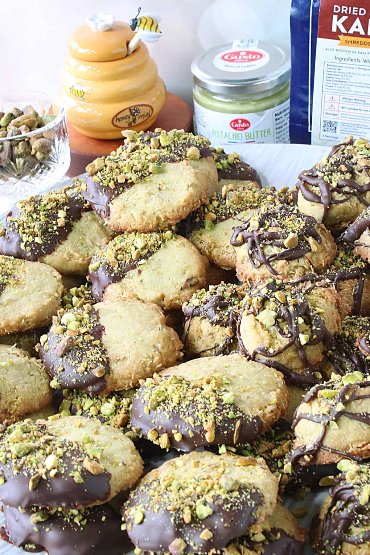 A jar of honey and some shelled pistachios behind a stack of Dubai chocolate bar cookies.