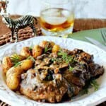 A white plate filled with steak with mushroom bourbon gravy and topped with fresh herbs.