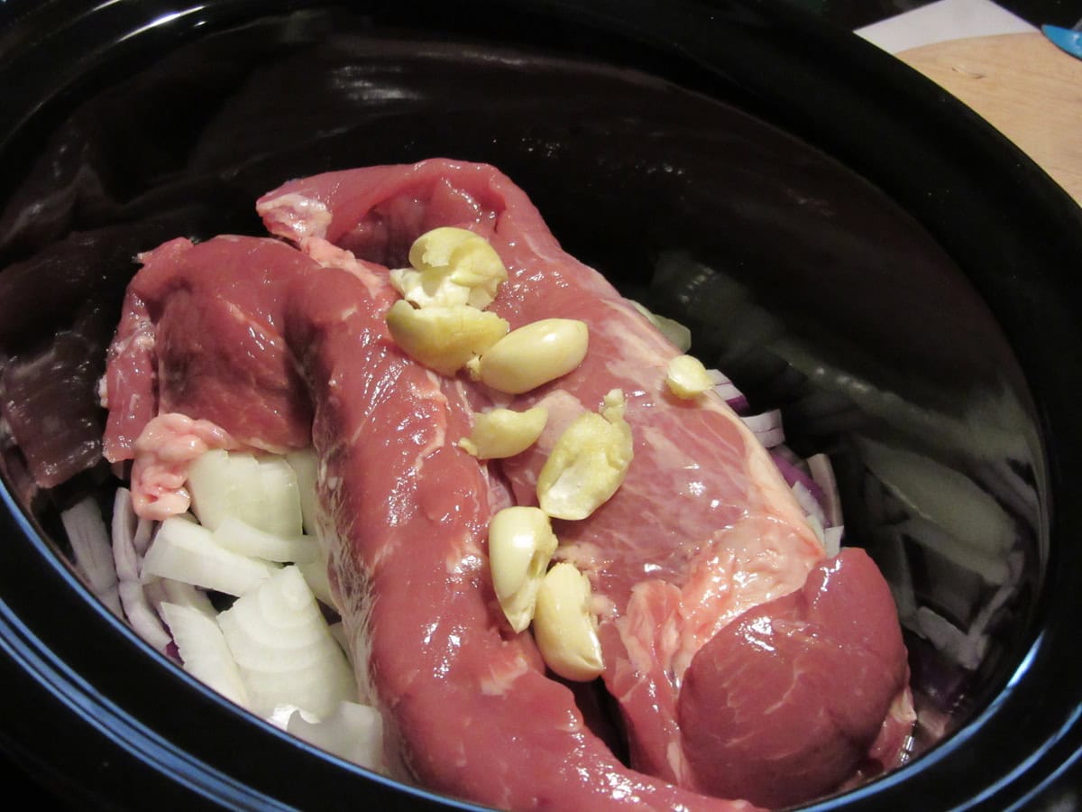 Garlic cloves, onions, and pork tenderloin in a slow cooker.