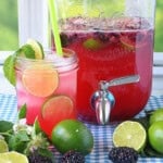 A mason jar glass filled with blackberry limeade and ice with fresh limes and blackberries in the foreground.