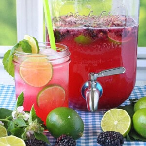 A mason jar glass filled with blackberry limeade and ice with fresh limes and blackberries in the foreground.