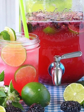 A mason jar glass filled with blackberry limeade and ice with fresh limes and blackberries in the foreground.