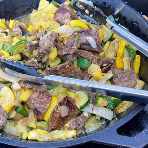A pair of cooking tongs in a skillet with cooked flank steak and squash.