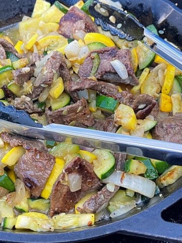 A pair of cooking tongs in a skillet with cooked flank steak and squash.