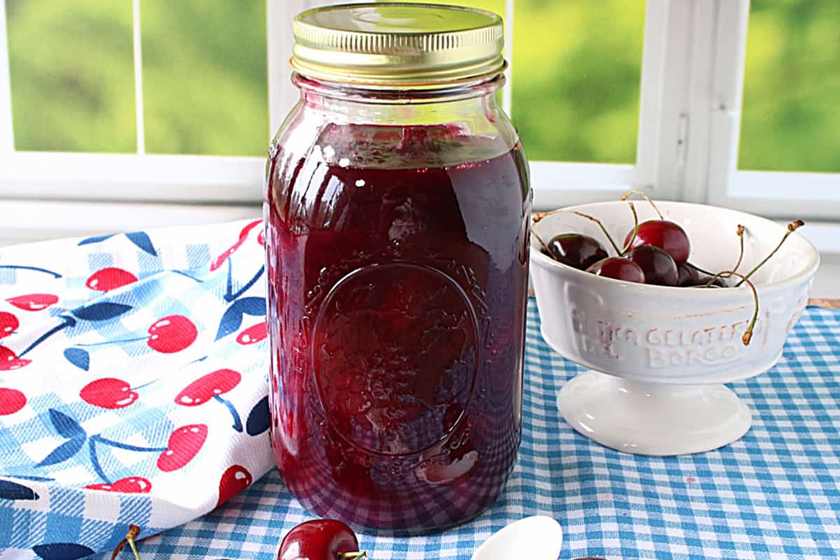 A blue and white checked table cloth with a jar of homemade cherry pie filling on top.