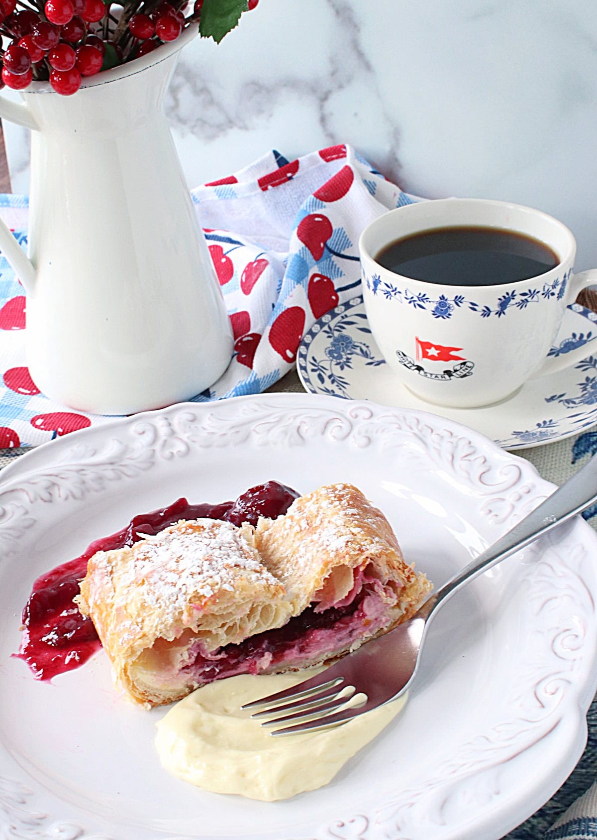 A cup of coffee in the background and a plate with some cherry strudel in the foreground.