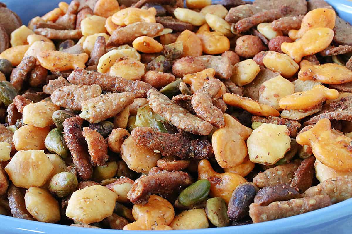 A snack mix closeup with sesame seed sticks, pistachios, rye crackers, and peanuts in a blue bowl.