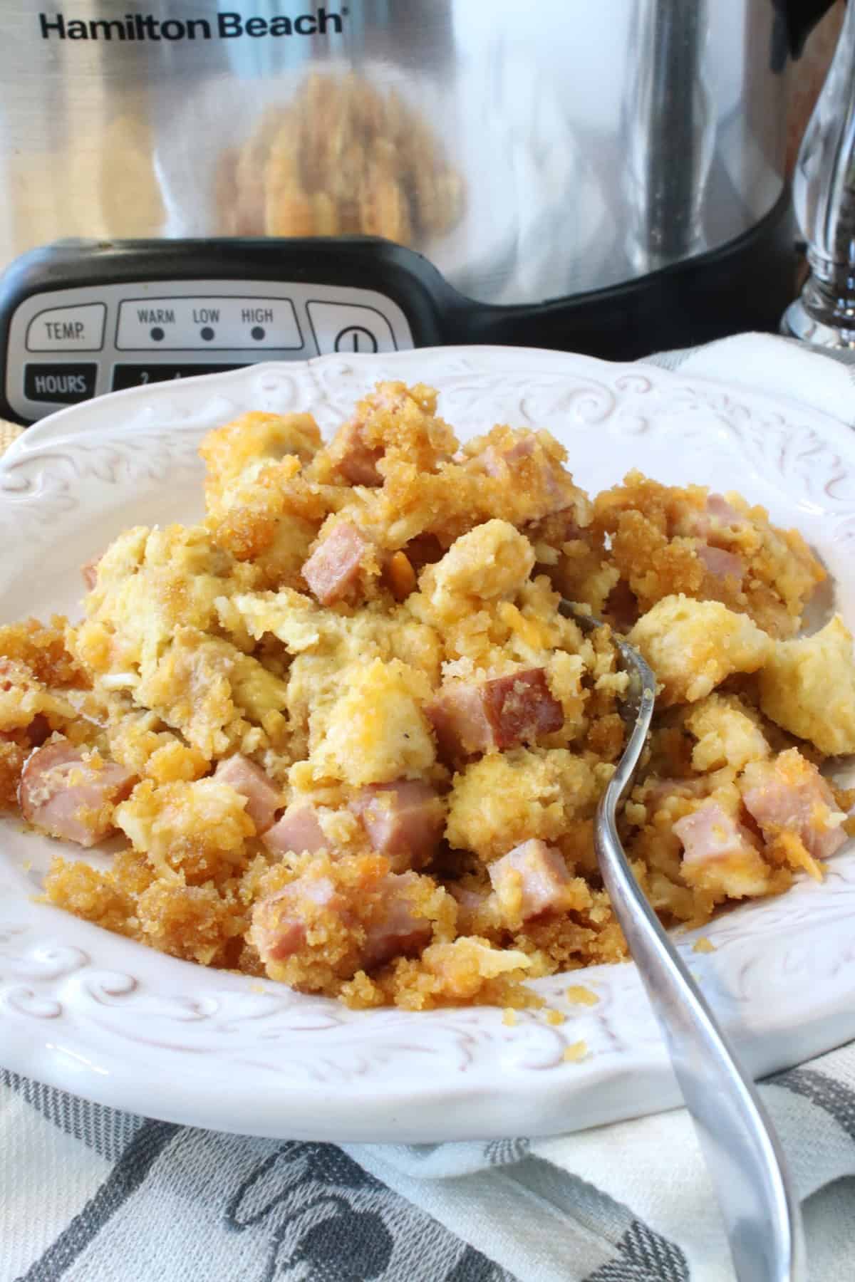 A bowl with a serving of slow cooker ham and cheese strata along with a slow cooker in the background.