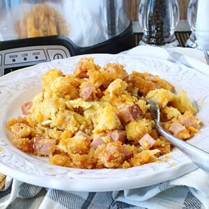 A bowl, spoon, and serving of ham and cheese strata with a slow cooker in the background.