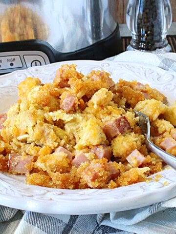 A bowl, spoon, and serving of ham and cheese strata with a slow cooker in the background.