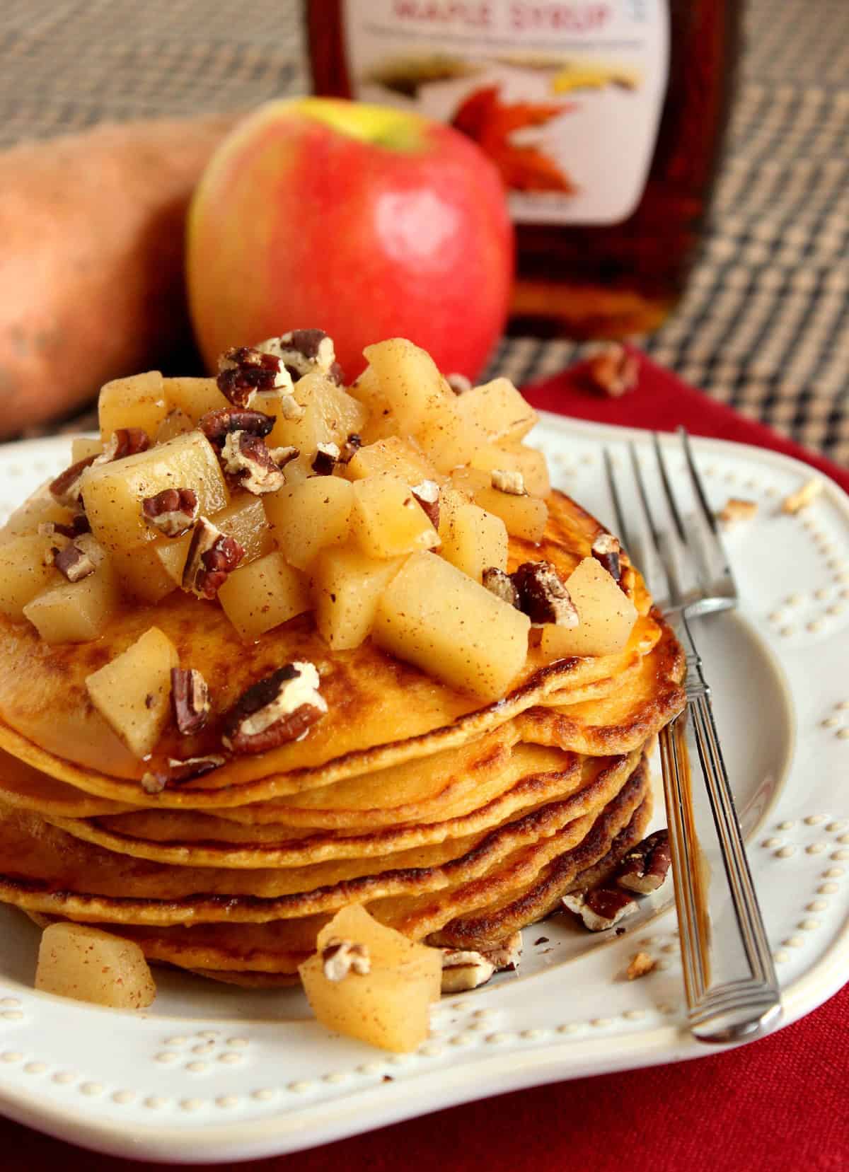 A whole apple in the background of a stack of sweet potato pancakes on a plate in the foreground.