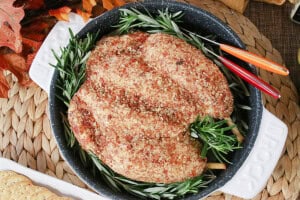 A realistic turkey cheeseball with rosemary, pecans, and bacon in a white roaster pan.