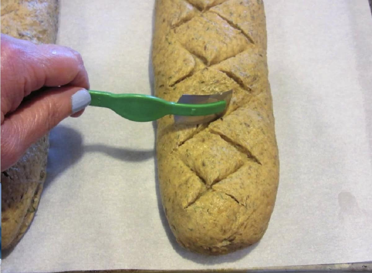 A hand with a bread lame slicing slits in rye bread dough.